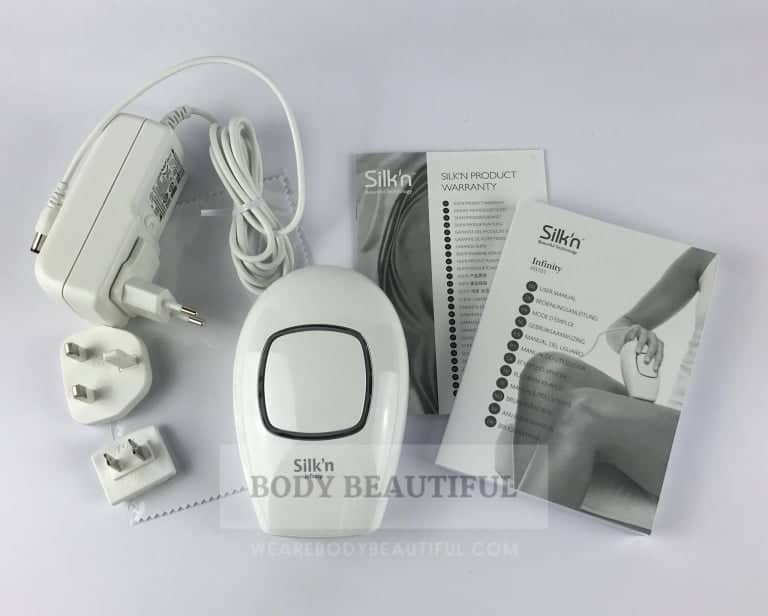 Top-down photo of the storage box contents on white background: power adaptor with UK, European and US plug adaptors, cleaning cloth in protective plastic, compact white Silk'n Infinity device, warranty card and multi-language user instruction booklet.